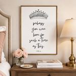 A framed wall art print with a black crown illustration and the quote 'perhaps you were born for such a time as this.' hangs above a wooden nightstand in a bedroom setting, with a lamp, books, flowers, and part of a bed visible.