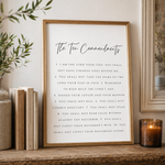 A framed print of The Ten Commandments displayed on a wooden shelf next to books and a lit candle. The print features elegant black script and serif text on a clean white background, enclosed in a thin gold frame.