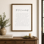 A framed 'The Ten Commandments' wall art print hangs on a light beige wall above a wooden console table. The print features elegant black text on a white background, enclosed in a natural wood frame.