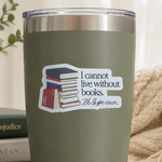 An olive green insulated tumbler with a clear lid sits on a wooden table in a cozy room setting. The tumbler features a white die-cut sticker with a stack of books and the quote, 'I cannot live without books.
