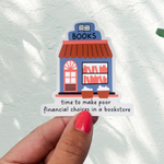 A hand with red nail polish holds a small die-cut sticker featuring an illustrated bookstore and the text 'time to make poor financial choices in a bookstore' against a white wall with leaf shadows.